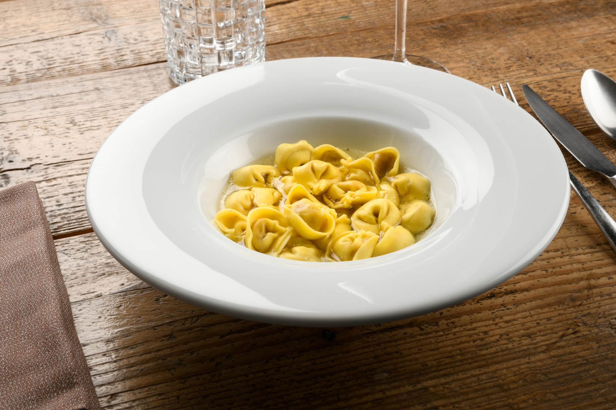 Piatti di tortellini in brodo su tavolo di legno.