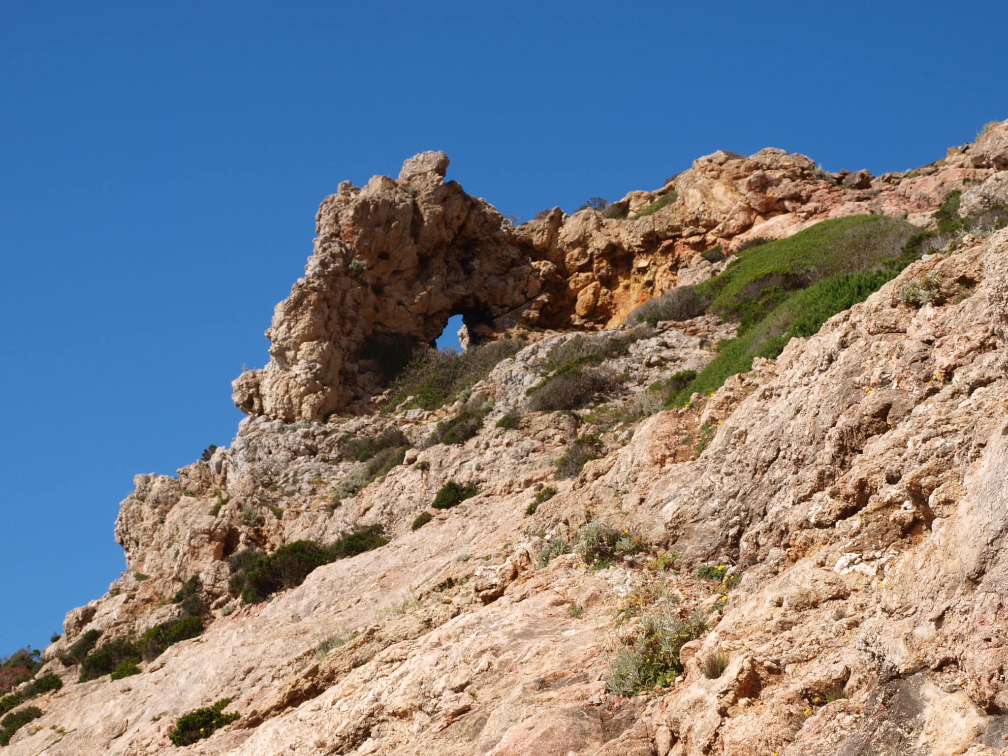 Scogliera rocciosa con arco naturale e cielo azzurro.