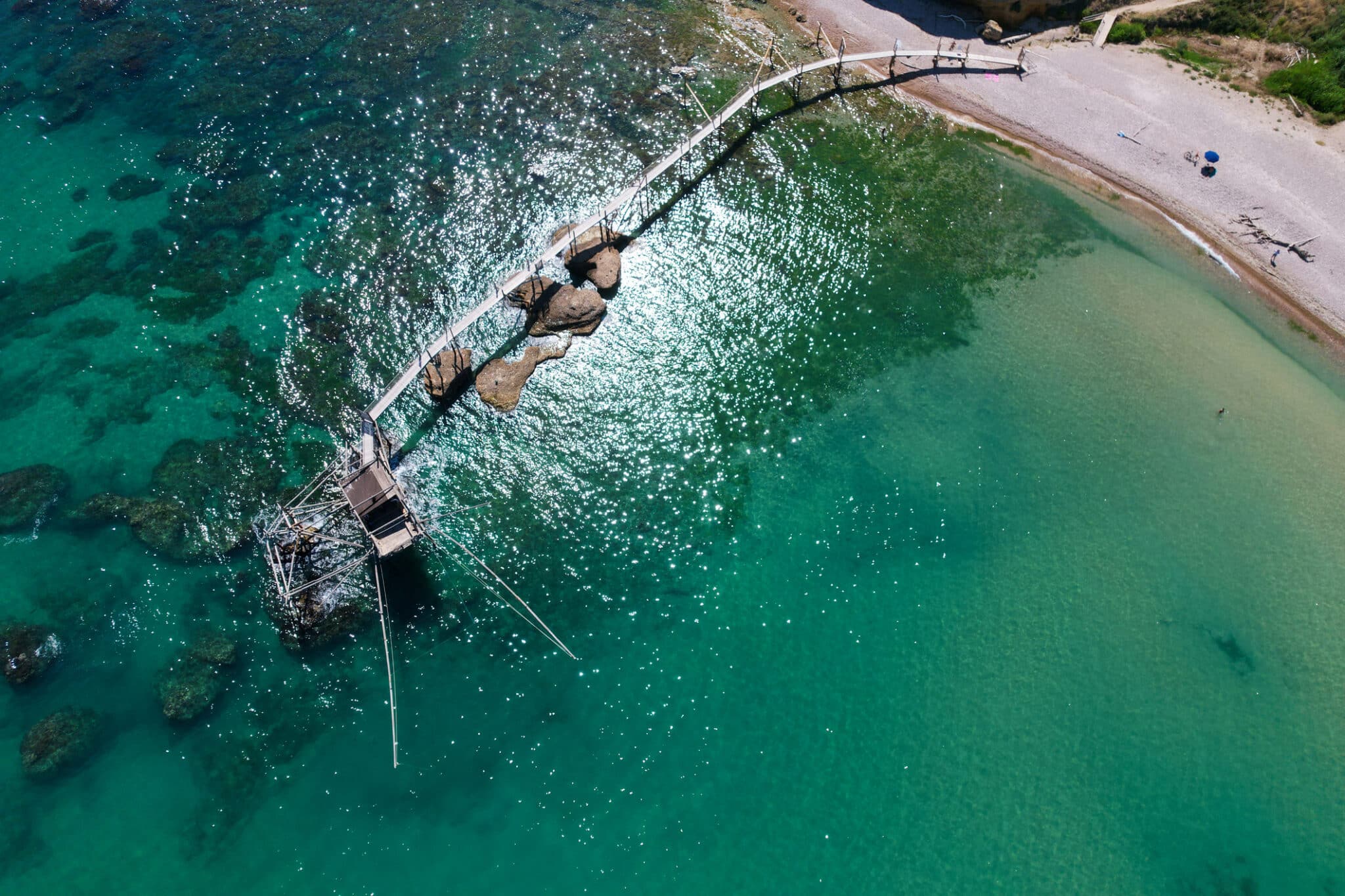 Ponte in legno su spiaggia e mare cristallino dall'alto.