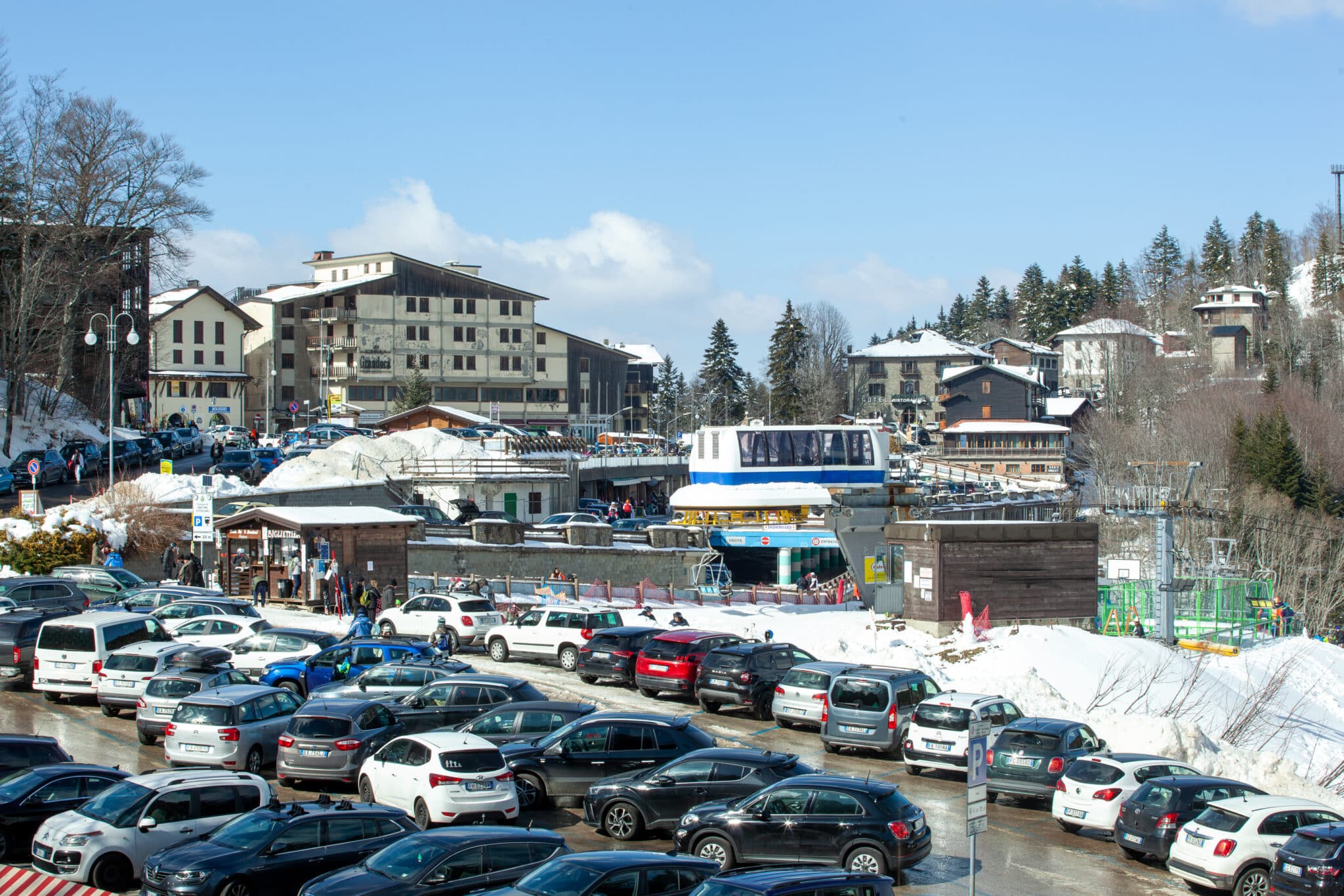 Parcheggio affollato in montagna con neve e impianti di risalita.