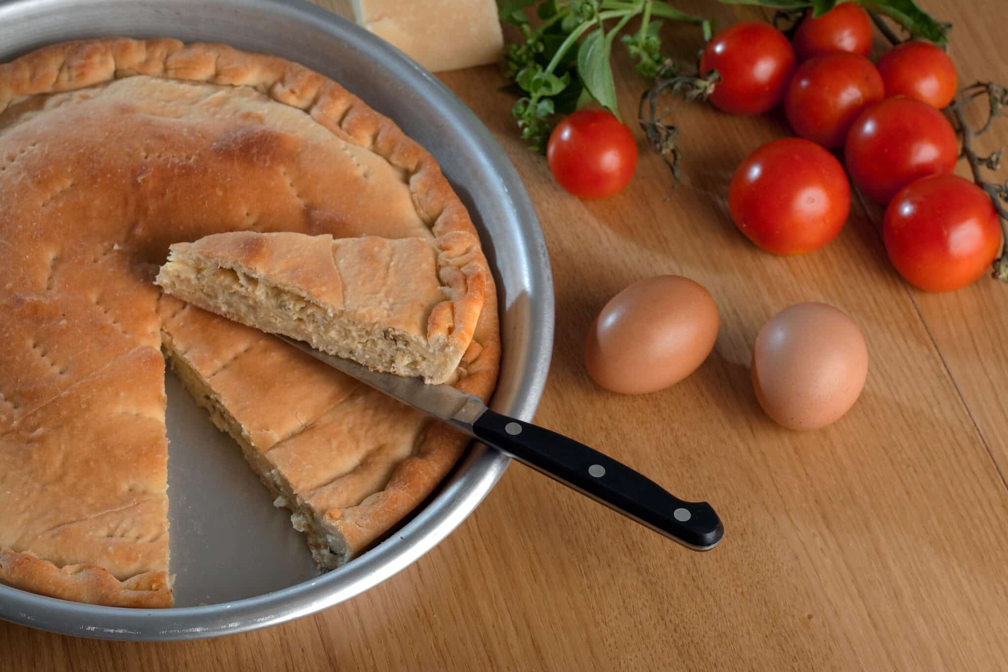 Ein Tiella-Kuchen aus Gaeta in Italien auf einem Holztisch mit Kirschtomaten, Basilikum und Eiern